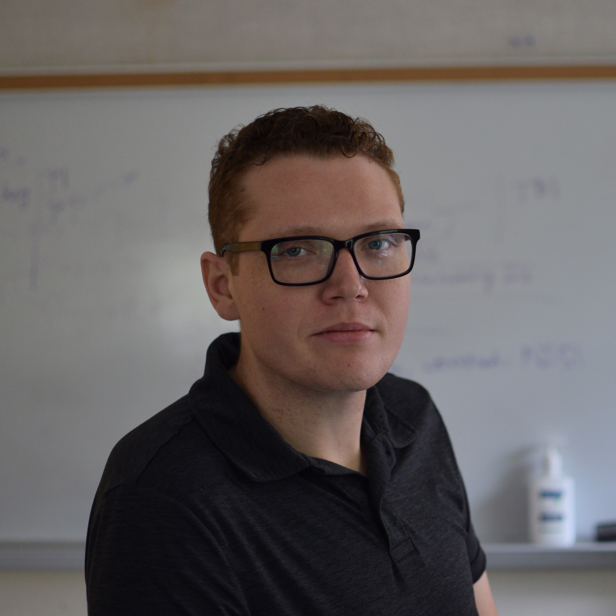 Myself sitting on a table with my short curly hair, nerdy glasses, and a dark grey polo shirt on. Behind me is a whiteboard with some diagrams written on it. I am looking directly into the camera with a slight smirk on my face.